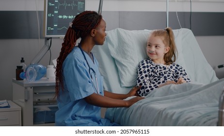 African American Pediatrician Nurse Sitting Beside Sick Child Giving High Five Discussing Healthcare Treatment During Recovery Examination In Hospital Ward. Little Kid Suffering Medicine Surgery