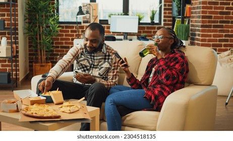 African american people using smartphones on couch, sitting together in front of television and browsing mobile phone apps. Modern partners enjoying leisure time with fast food meal. - Powered by Shutterstock