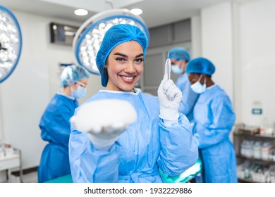 African American Plastic Surgeon Woman Holding Silicon Breast Implants And A Scalpel In Surgery Room Interior. Cosmetic Surgery Concept