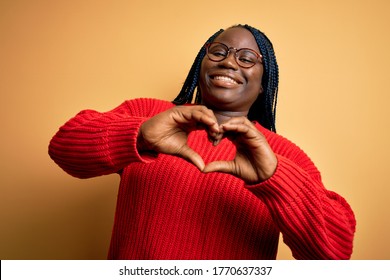 African american plus size woman with braids wearing casual sweater over yellow background smiling in love doing heart symbol shape with hands. Romantic concept.