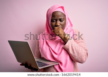 African american plus size woman wearing muslim hijab using laptop over pink background feeling unwell and coughing as symptom for cold or bronchitis. Health care concept.