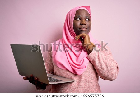 African american plus size woman wearing muslim hijab using laptop over pink background with hand on chin thinking about question, pensive expression. Smiling with thoughtful face. Doubt concept.
