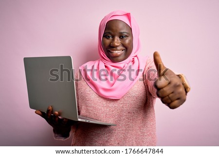 African american plus size woman wearing muslim hijab using laptop over pink background approving doing positive gesture with hand, thumbs up smiling and happy for success. Winner gesture.