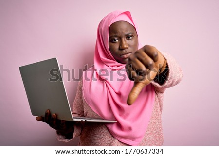African american plus size woman wearing muslim hijab using laptop over pink background looking unhappy and angry showing rejection and negative with thumbs down gesture. Bad expression.