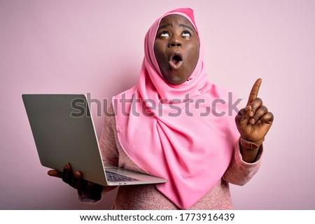 African american plus size woman wearing muslim hijab using laptop over pink background amazed and surprised looking up and pointing with fingers and raised arms.