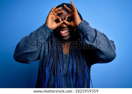 African american plus size woman with braids wearing casual sweater over blue background doing ok gesture like binoculars sticking tongue out, eyes looking through fingers. Crazy expression.