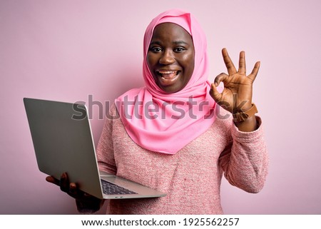 African american plus size woman wearing muslim hijab using laptop over pink background smiling positive doing ok sign with hand and fingers. Successful expression.
