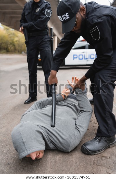 african american\
police officer with truncheon arresting hooded offender lying on\
street on blurred\
background