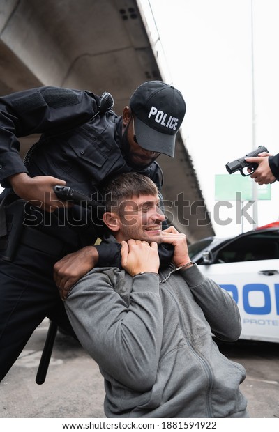african american policeman choking handcuffed\
offender while aiming with pistol near colleague on blurred\
background outdoors