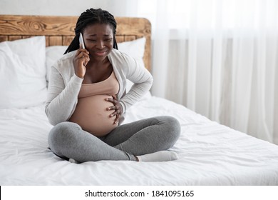 African American Pregnant Lady Feeling Pain In Belly, Calling Doctor On Cellphone While Sitting On Bed At Home, Suffering From Abdominal Ache, Black Expectant Woman Having Labor Cramps, Copy Space