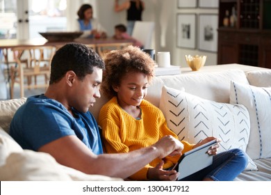 African American pre-teen girl sitting on sofa in the living room using tablet computer with her father, mother and toddler sitting at a table in the background, selective focus - Powered by Shutterstock