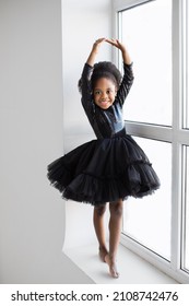 African american pretty ballerina in black dress dancing barefoot on windowsill. Isolated over white studio background. Grace of little lady.