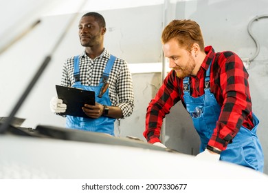 African American and red-haired craftsmen are standing in overalls and looking at the open hood of the car thoughtfully - Powered by Shutterstock