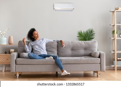 African american relaxed woman sitting on comfortable couch in living room at modern home holds air conditioner remote control enjoying breathing fresh cool air at summer or warm air at winter season - Shutterstock ID 1332865517