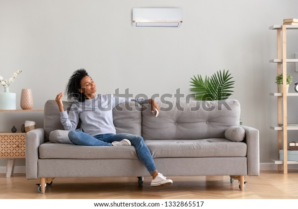 African american relaxed woman sitting on\
comfortable couch in living room at modern home holds air\
conditioner remote control enjoying breathing fresh cool air at\
summer or warm air at winter\
season