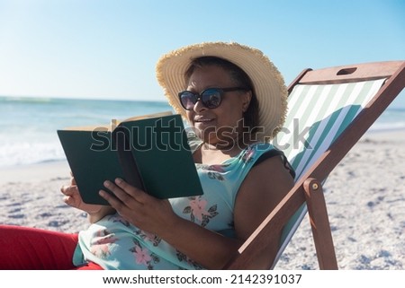 African american retired senior woman reading book while sitting on folding chair at beach. unaltered, hobbies, active lifestyle, enjoyment and holiday concept.