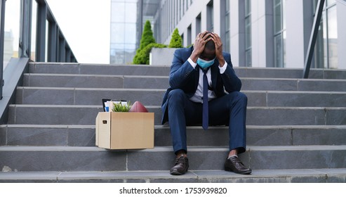 African American Sad Businessman In Medical Mask Sitting On Stairs Outdoor With Box Of Stuff As Lost Business. Male Office Worker In Despair Lost Job. Unemployment Rate Growing Due Pandemic. Fired Man