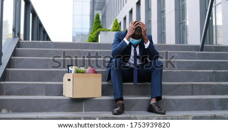 African American sad businessman in medical mask sitting on stairs outdoor with box of stuff as lost business. Male office worker in despair lost job. Unemployment rate growing due pandemic. Fired man