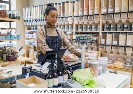 African american saleswoman or small business owner weighs some kraft paper package on scales at sustainable small local business. Young afro woman at cashdesk with scales in local zero-waste grocery 