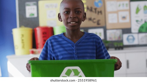 African american schoolboy smiling, holding recycling bin, standing in classroom. children at primary school in summer. - Powered by Shutterstock