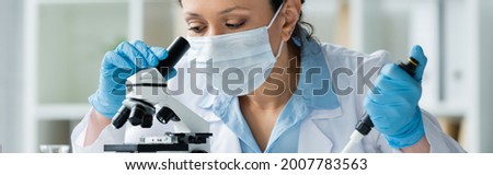 African american scientist in medical mask holding electronic pipette near microscope, banner