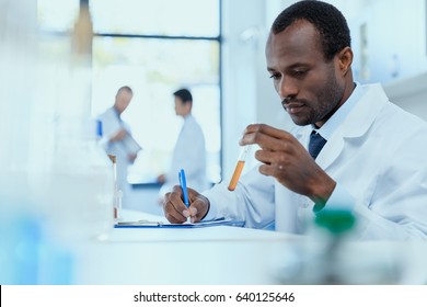 African american scientist in white coat holding and examining test tube with reagent, laboratory researcher concept
