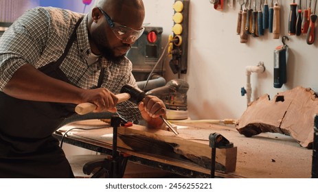 African american sculptor shaping raw timber using chisel and hammer in carpentry shop, creating wood art, wearing safety glasses. Artist making wood sculptures, engraving lumber with tools, camera A - Powered by Shutterstock