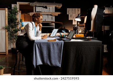 African american seamstress in atelier shop workspace surrounded by sewing instruments looking online for design inspirations, inspecting sketch drawings of upcoming sartorial collection - Powered by Shutterstock