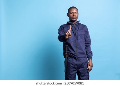 African american security officer raising a finger in front of the camera, showing a restriction symbol against blue background. Man working as a patrol agent presents a stop sign in studio. - Powered by Shutterstock