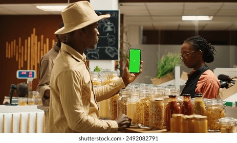 African american seller holding smartphone app with greenscreen, presenting isolated mockup template in local supermarket. Vegan merchant uses chromakey layout at organic grocery store. Camera 1. - Powered by Shutterstock