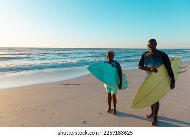 African american senior couple carrying surfboards while walking at sandy beach against sky. Copy space, water sports, recreation, unaltered, vacation, sunset, together, retirement, enjoyment, nature. - Powered by Shutterstock