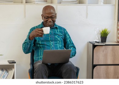 African American senior man with a coffee mug and using a smartphone while working with a laptop at home. - Powered by Shutterstock