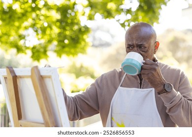 African american senior man drinking coffee and painting on canvas on sunny terrace. Lifestyle, retirement, senior lifestyle, nature, creativity and domestic life, unaltered. - Powered by Shutterstock