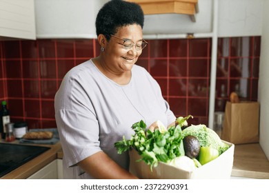 African american senior overweight female holding big carton box with green fresh organic vegetables standing in her kitchen, going to cook delicious and healthy dinner being on diet - Powered by Shutterstock