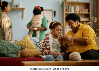 African American senior woman teaching child knitting skills while sitting on couch together during family gathering in living room