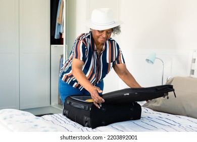 African American Senior Woman Wearing Hat Packing Suitcase In Bedroom. Travel Preparation During Covid 19 Pandemic.