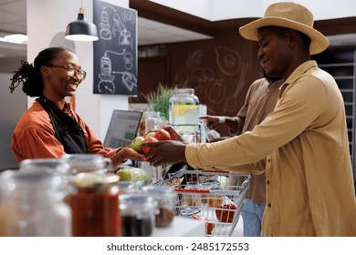 African American shopkeeper of an eco friendly business with fresh organic items helping clients. Focus on a black man presenting fruits to a female employee at the checkout desk. side view shot. - Powered by Shutterstock