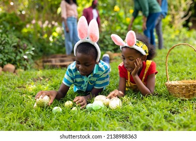 African American Siblings In Bunny Ears Arranging Easter Eggs On Grass While Family In Background. Unaltered, Lifestyle, Easter Day, Celebration, Childhood, Cultures And Holiday Concept.