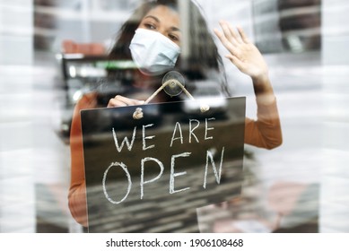 African American Small Business Owner In Medical Mask Holds A Signboard WE ARE OPEN At The Entrance To A Restaurant, Bar, Cafe Or Shop, And Waiting For Visitors