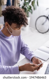 African American Small Business Owner In Protective Mask Using Smartphone At Working Table