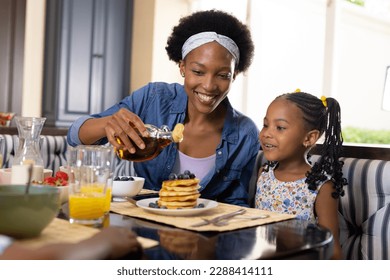 African american smiling mother pouring honey on pancakes while having breakfast with daughter. Unaltered, family, togetherness, childhood, food, fresh, dining table and home concept. - Powered by Shutterstock
