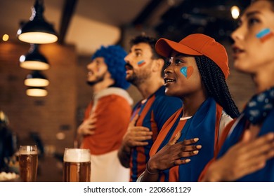 African American soccer fan and her friends with hand on heart singing national anthem while watching sports championship in bar. - Powered by Shutterstock