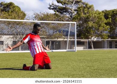 African American soccer player celebrating on soccer field, looking strong and fit, copy space. Green grass and goalpost in background with trees and buildings nearby, unaltered - Powered by Shutterstock