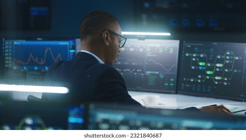 African American software engineer sits in front of computer and big digital screens with data server and blockchain network, works in modern monitoring control room with real-time analysis charts.