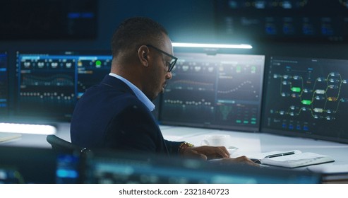 African American software engineer sits in front of computer and big digital screens with data server and blockchain network, works in modern monitoring control room with real-time analysis charts. - Shutterstock ID 2321840723