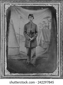African American soldier posed in front of quaint studio backdrop of tents in a military camp with a prominent U.S. flag. The soldier is standing on the dirt ground, by a traveling photographer. 1865.