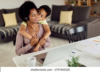 African American son embracing and kissing his mother who is working on a computer at home. 