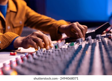 African american sound engineer twisting knobs to edit music in studio, operating control panel board desk with motorized faders and buttons. Skilled producer creating records with gear. Close up.