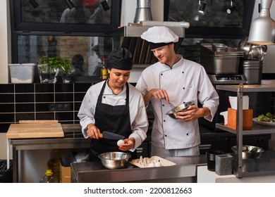 African American Sous Chef Holding Knife Near Raw Egg Above Bowl While Cooking Near Colleague In Kitchen