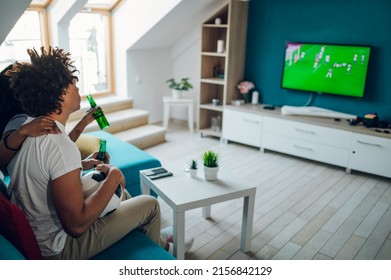 African American Sports Fan Couple Watching Important Soccer Match On TV While Drinking Beer And Cheering At Home. Guy Holding A Soccer Ball. Cheering For A Favorite Team.